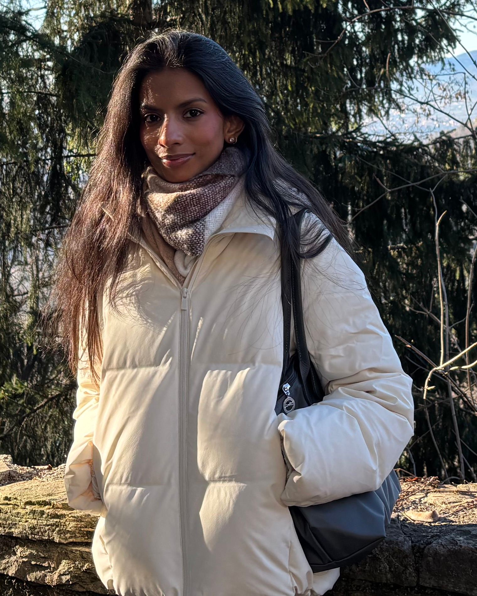 Rhythm Gangwar, standing outdoors in a cream puffer jacket and a patterned wool scarf, a wooded hillside and valley behind her.